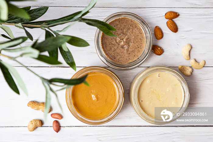 The modern concept of wellness and veganism. Jars of almond, cashew and peanut butter on a white wooden table with an olive branch. Organic, homemade nut butter for a healthy breakfast.