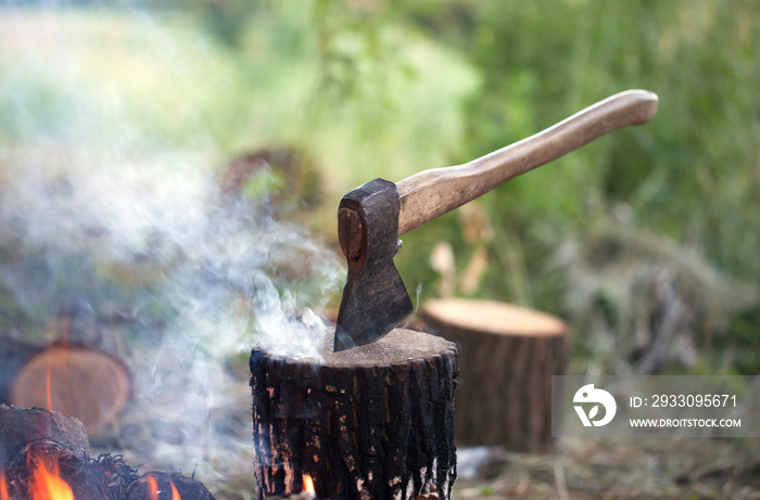 Axe in tree stump and campfire with smoke in summer forest