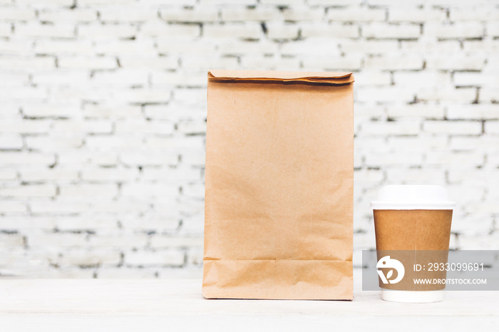 Cup of coffee and blank paper bag with copy space for your brand on table against white brick wall background