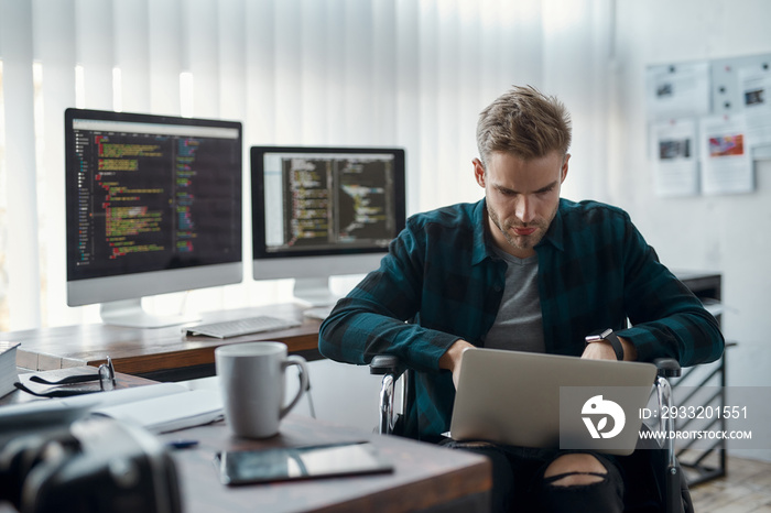 Portrait of young focused male web developer in a wheelchair writing program code on his laptop at h