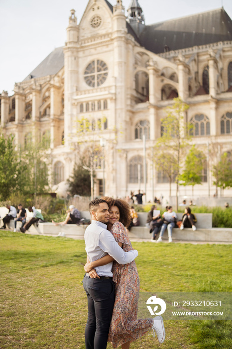 Side view of happy couple embracing while standing on grassy field against building in park
