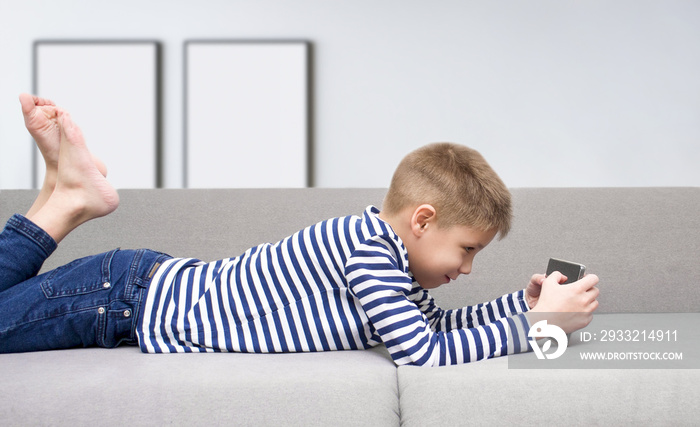 The boy lies on the couch and looks into the tablet. The child plays or watches a cartoon on a table