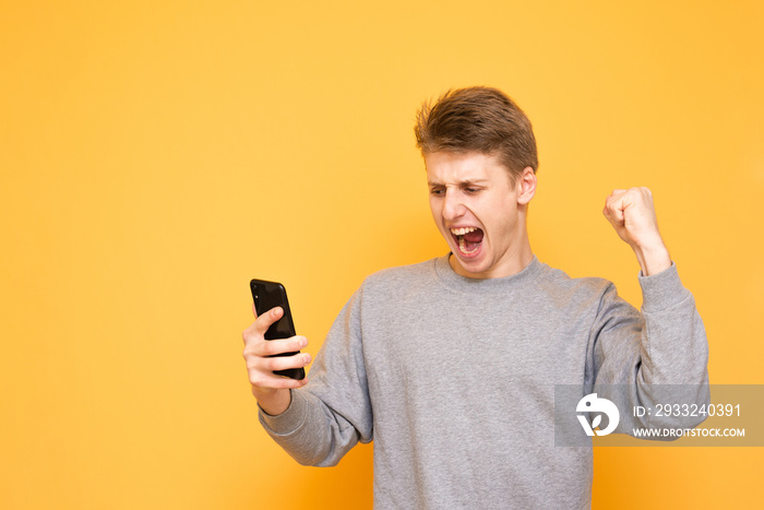 Expressive guy in casual clothing looks at the smartphone screen and rejoices, hands up on a yellow 