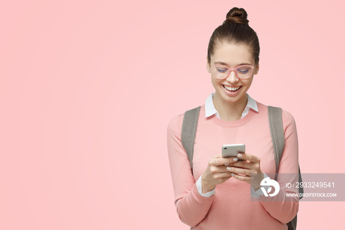 Close-up portrait of young female student looking excited at display of smartphone, smiling and laug
