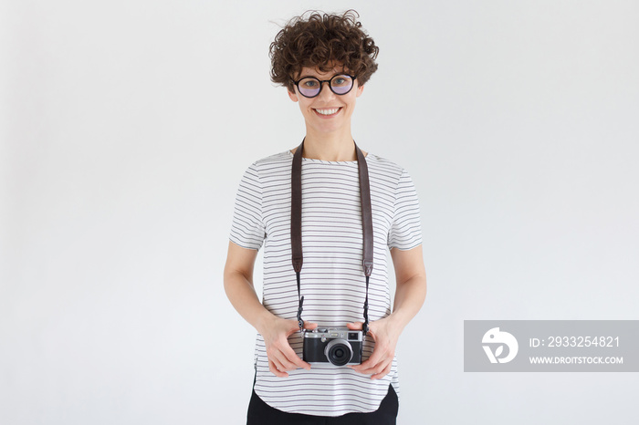 Portrait of smiling female photographer holding her camera, standing isolated on gray background