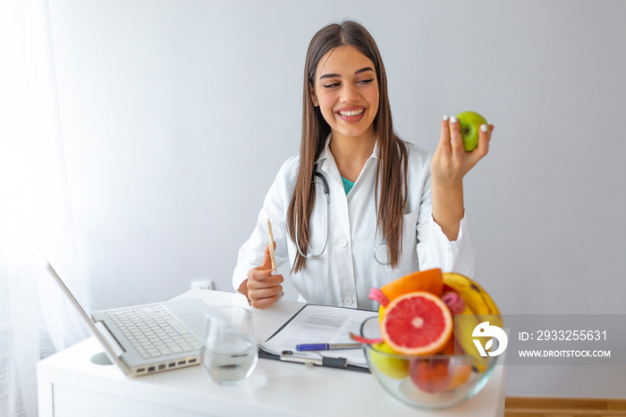 Smiling nutritionist in her office, she is holding a green apple and showing healthy vegetables and 