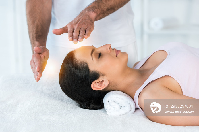 cropped view of healer putting hands near head of woman with closed eyes lying on massage table