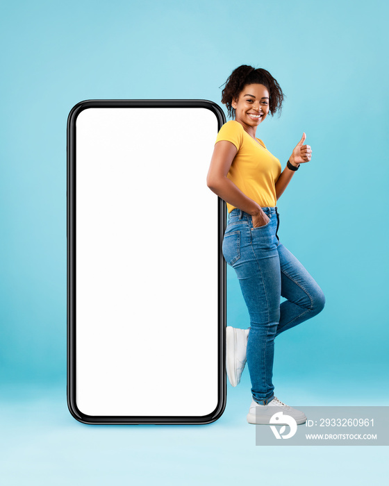 Pretty black lady standing near big smartphone with blank screen, gesturing thumb up on blue backgro