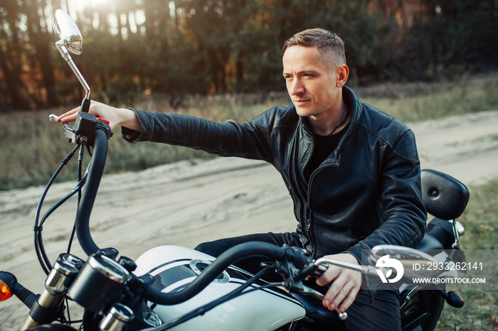 Handsome rider guy in black biker jacket on classic style cafe racer motorcycle at sunset. Bike cust