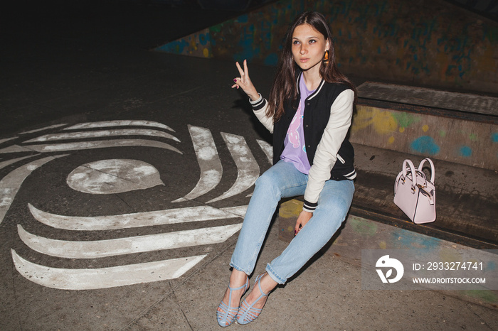 young stylish hipster woman, swag outfit, jeans, cool accessories, sitting on ground with purse