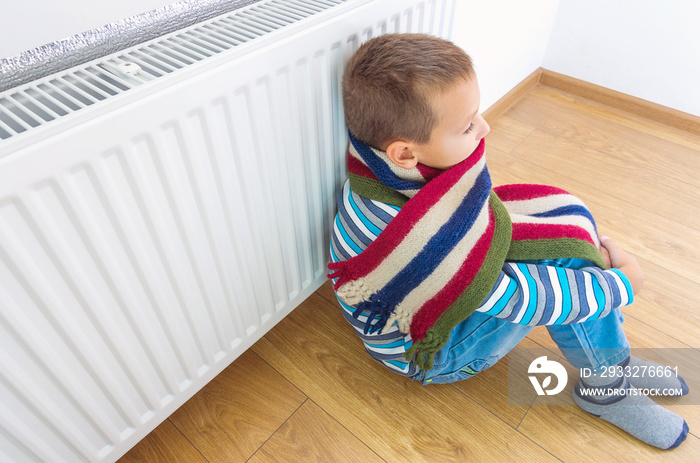 A young boy in a warm woolen sweater and scarf warms himself near a heating radiator at home. The sy