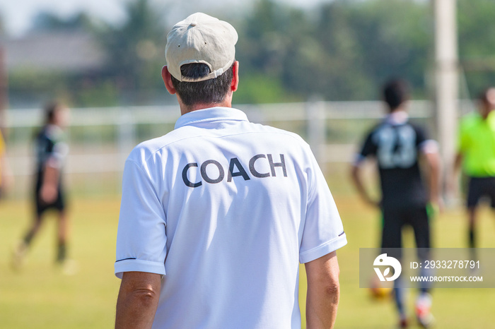 Back of male football coach wearing white COACH shirt at an outdoor sport field coaching his team du