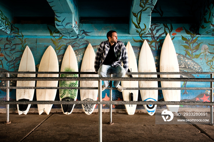 Thoughtful man sitting on railing against surfboards in graffiti tunnel