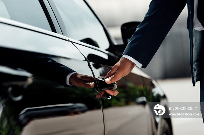 cropped view of african american businessman in suit opening car door at sunny day