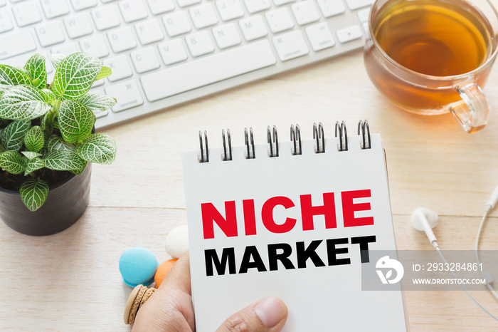 Man holding NICHE MARKET message on book and keyboard with a hot cup of tea, macaroon on the table. 