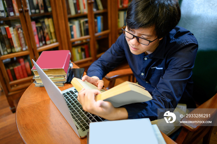 Asian man university reading book and using laptop computer nearby vintage bookcase or bookshelf in 