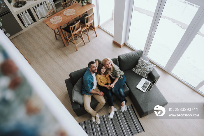 Loving parents sitting on couch with laptop and hugging their daughter