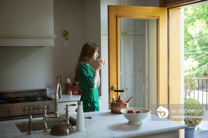 Side view of thoughtful woman holding tea cup while standing in kitchen at home