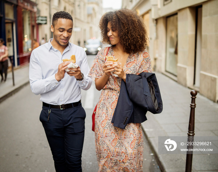 Happy couple eating food while walking on street amidst buildings in city