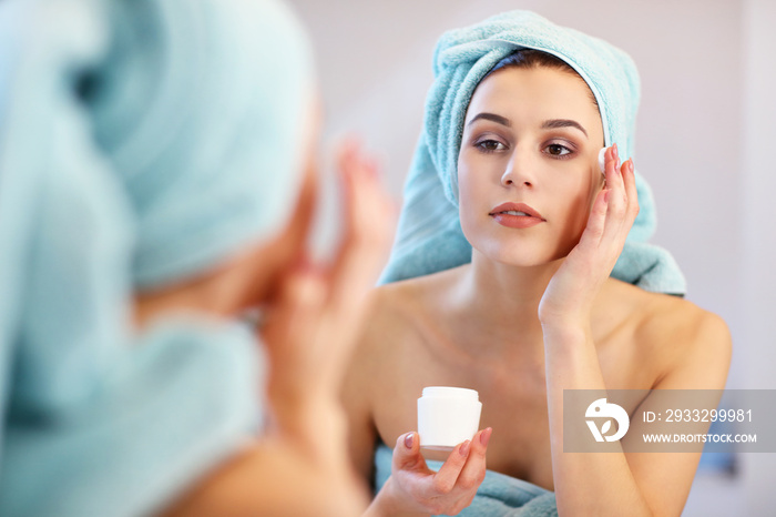 Young woman standing in bathroom and applying face cream in the morning