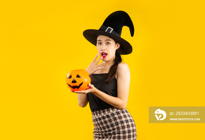 Young asian woman in black tank top wearing witch hat holding pumpkin lantern posing on yellow backg