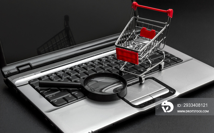 Shopping cart with magnifying glass and laptop on black background. Find purchase online.