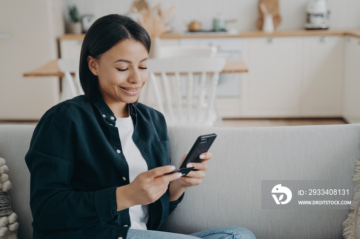 Woman holding bank credit card, smartphone makes successful cashless payment sitting on sofa at home