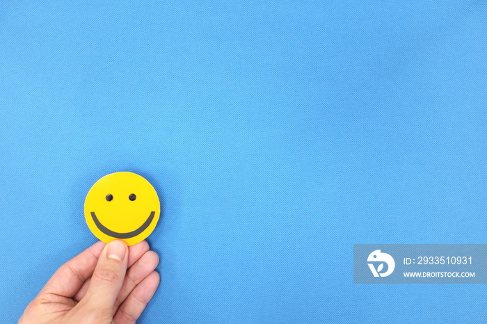 Happiness and positivity concept. Hand holding yellow smiling face in blue background with copy space.