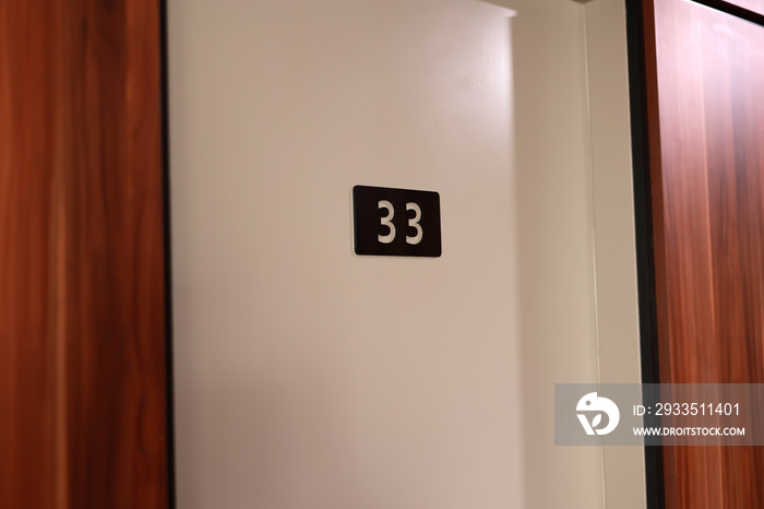 Hotel room 33 number mounted on a white wall. White number on black board by wooden doors in corridor hall