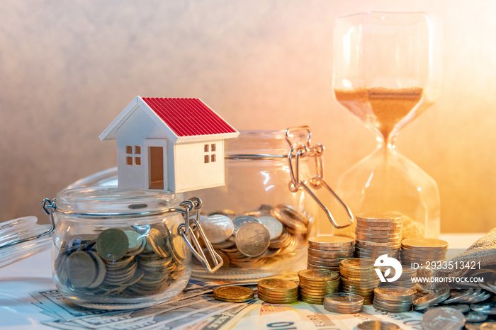 Saving money for retirement concept. Real estate or property investment. Home mortgage loan rate. Coin stack, money bag and currency glass jars on banknotes with hourglasses, house model on the table.