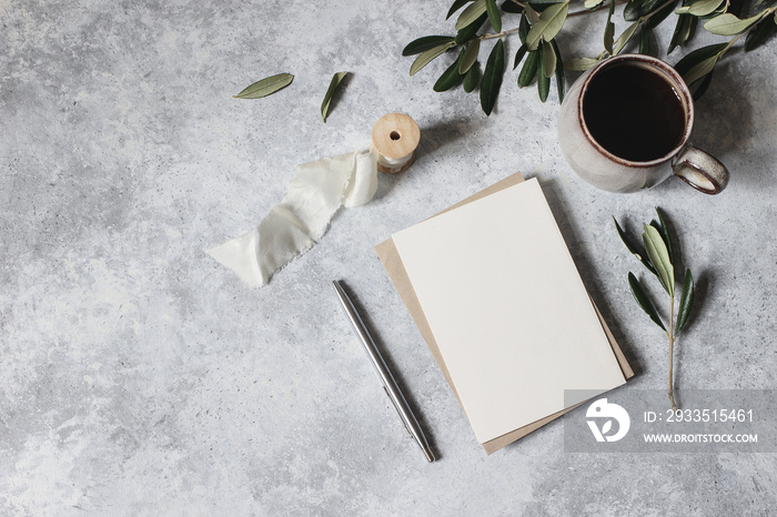 Moody feminine wedding stationery mock-up scene. Blank greeting card, cup of coffee, silver pen with olive leaves, branches. Grunge table background. Flat lay, top view. Mediterranean breakfast.