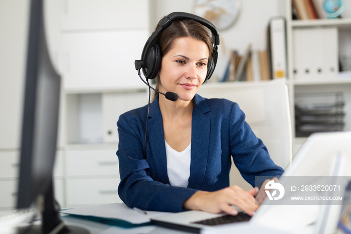 Portrait of positive woman call center operator receiving calls and using laptop in office