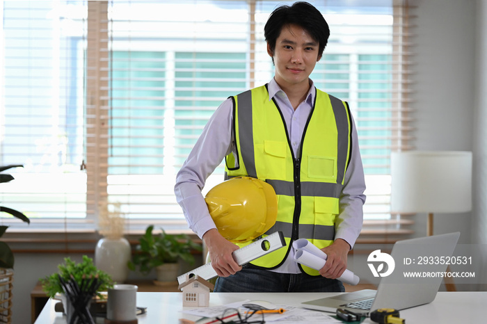 Professional construction site engineer in uniform holding safety helmet and standing at office.