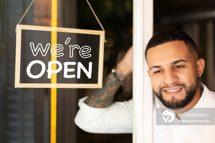 Smiling young Latin American man at the entrance of a small business next to a sign with the text We’re Open.