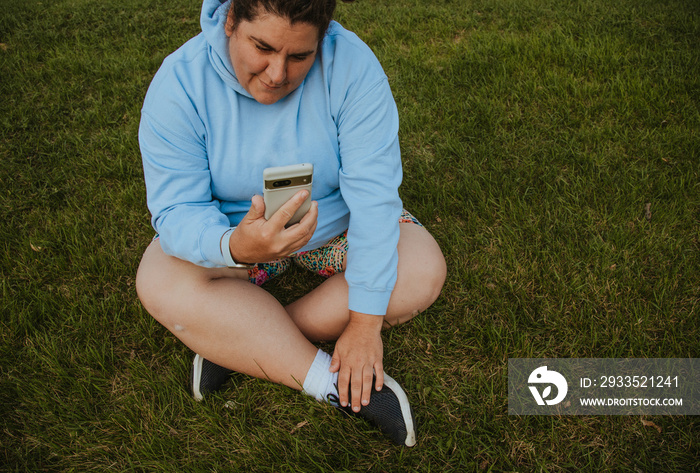 plus size woman sitting on grass wearing hoodie looking at phone