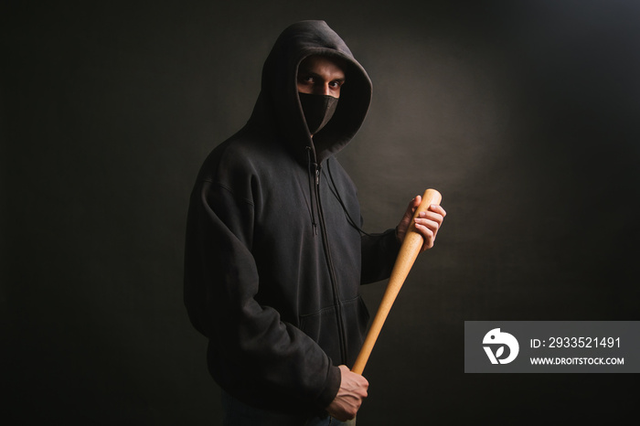 Bandit in a black sweatshirt and a medical mask. Dangerous man with a baseball bat ready to fight.