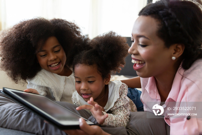 Happy afro family. Smiling African American mother with two little daughter looking at the digital tablet while lying on sofa at home. people, family, technology and education concept