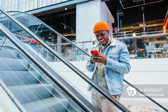 Emotional happy African-American guy in stylish outfit looks at mobile phone going up escalator in shopping mall close view