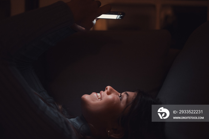 One beautiful young woman smiling and using phone at home at late night on the sofa. Happy female teenager chatting with friends surfing the net online. Enjoying technology and internet.