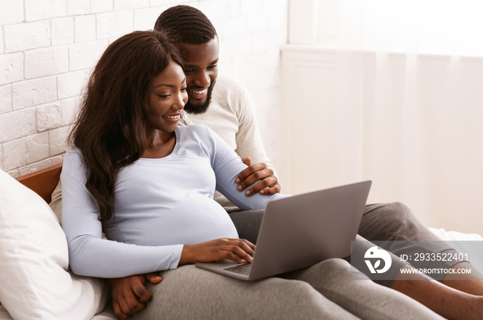 Pregnant woman and her husband using laptop, laying on bed