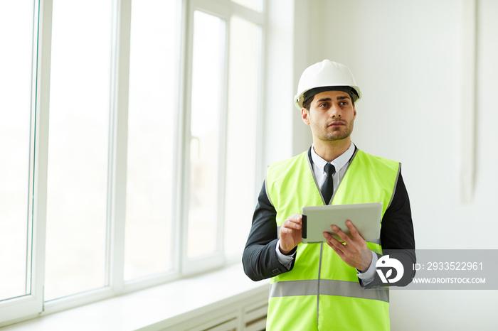 Waist up portrait of Middle-Eastern engineer wearing hardhat posing on construction site holding tablet, copy space