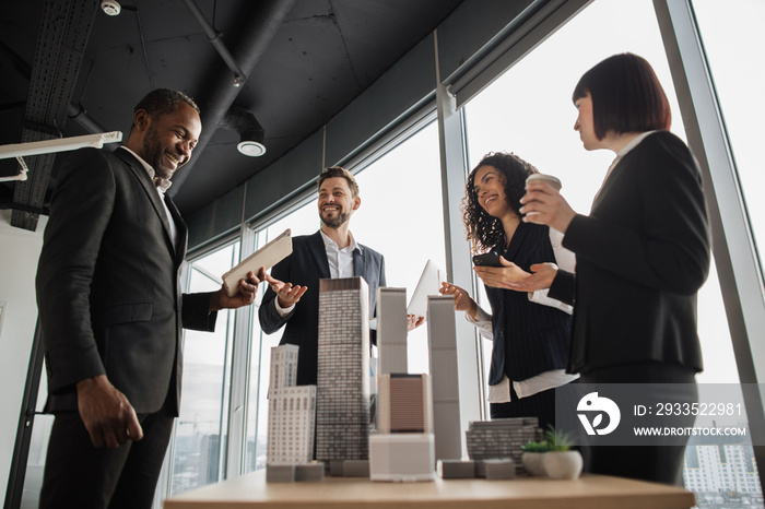 Multiethnic real estate agents, standing near table with 3d city model in office with panoramic city view, while handsome male project manager presenting them report about company’s activity on laptop