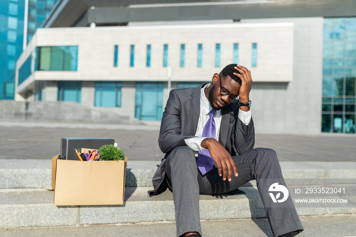 Unemployment concept. Disappointed black businessman sitting with box of personal stuff on stairs against office center