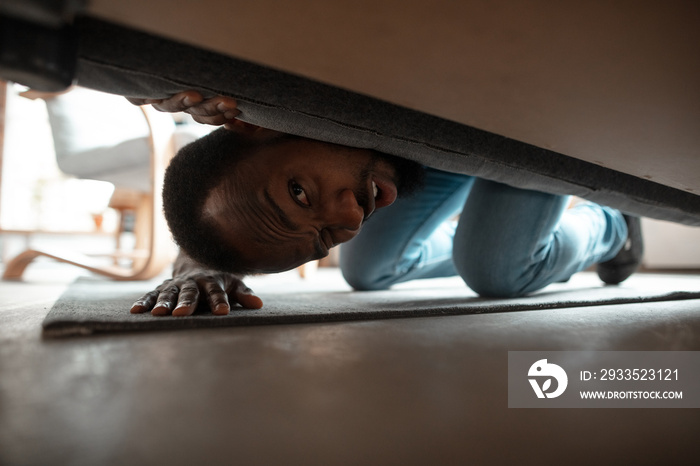 Under his bed in bedroom. African-american man looking for job in unusual places at his home. Crazy, funny way to find career and going up. Concept of crisis, unemployment, finance, business.