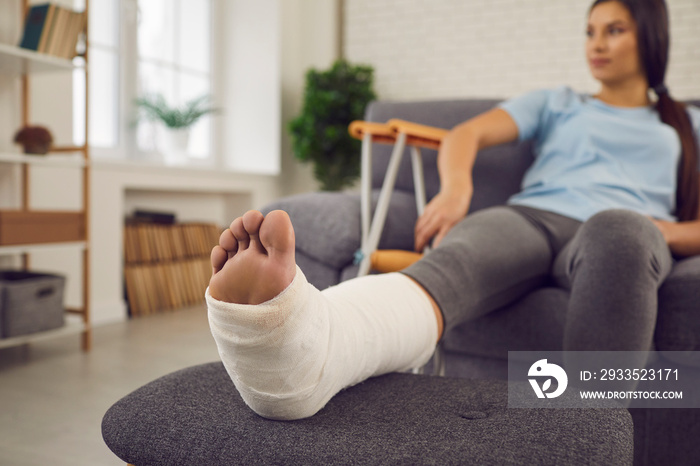 Concept of rehabilitation of people after serious physical accident injury. Female patient with broken leg in plaster cast sitting on sofa. Young woman with foot bone fracture resting on couch at home