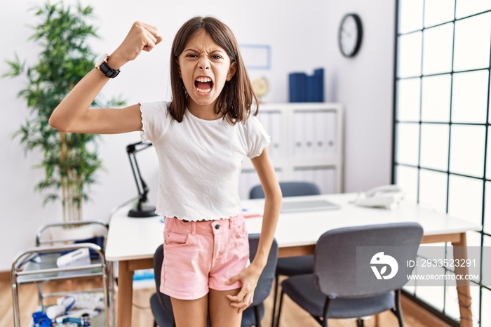 Young hispanic girl standing at pediatrician clinic angry and mad raising fist frustrated and furious while shouting with anger. rage and aggressive concept.