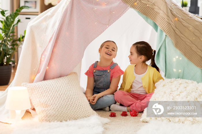 childhood and hygge concept - happy little girls playing tea party with toy crockery in kids tent at home