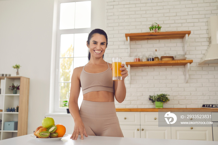 Happy young fitness woman athete in sportswear standing and drinking fresh orange juice from glass before or after workout at home. Active healthy lifestyle, clean eating concept