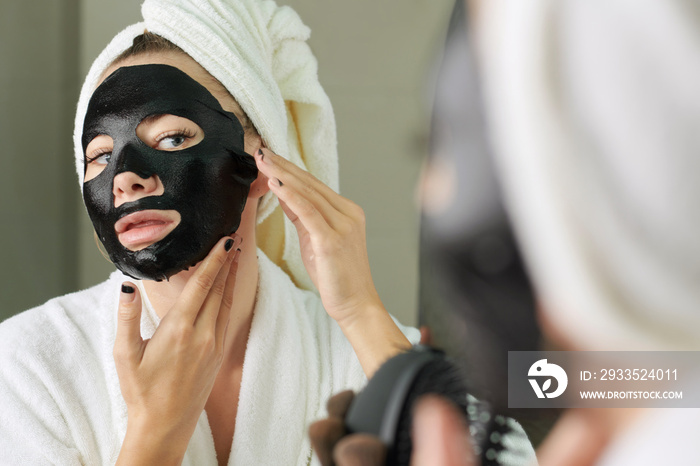 Woman applying black sheet mask on face after taking morning shower