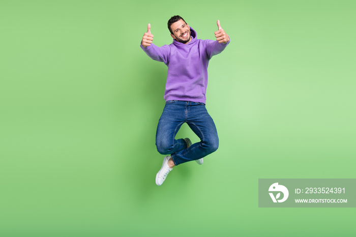 Full size photo of cool young brunet guy jump show thumb up wear hoodie jeans sneakers isolated on green background